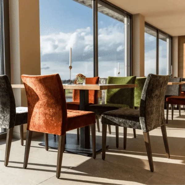 Sillas de terciopelo en tonos naranja, verde y gris en restaurante con grandes ventanales.