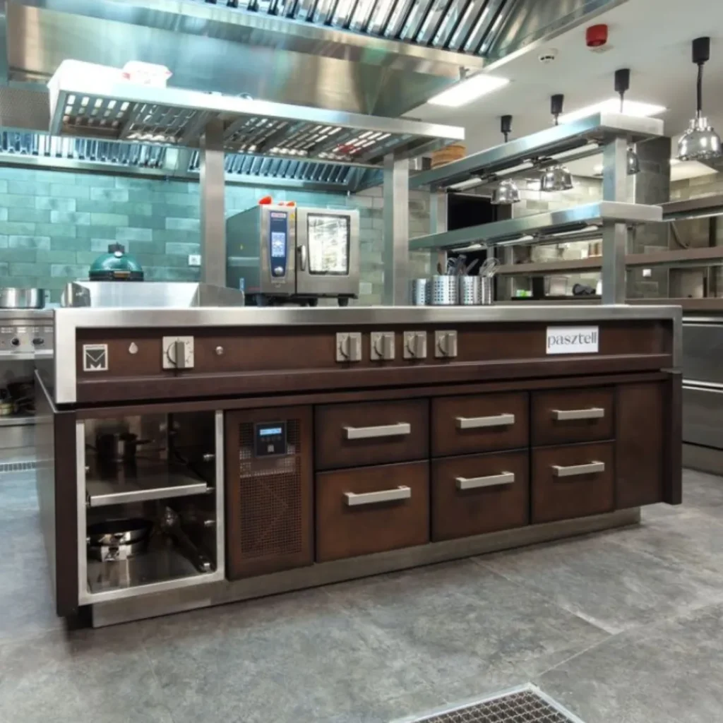 Estación de cocina Pasztell con encimera de acero inoxidable y frente de madera oscura, rodeada de equipo profesional.