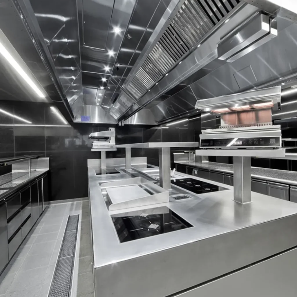 Estación de emplatado en cocina profesional con amplia encimera de acero inoxidable, lámparas de calor suspendidas y campana extractora.
