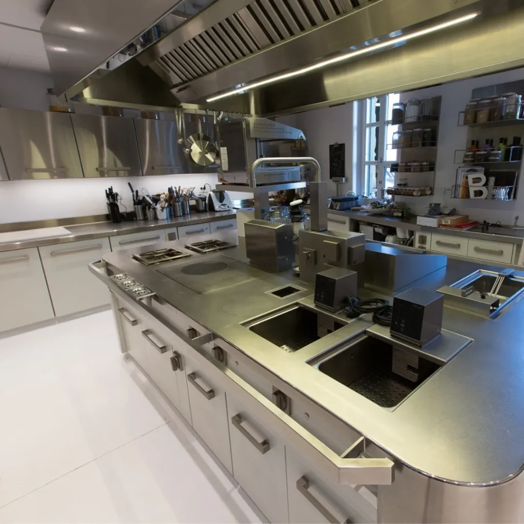 Estación de cocina profesional con encimera de acero inoxidable, fregaderos empotrados y estanterías con utensilios al fondo.