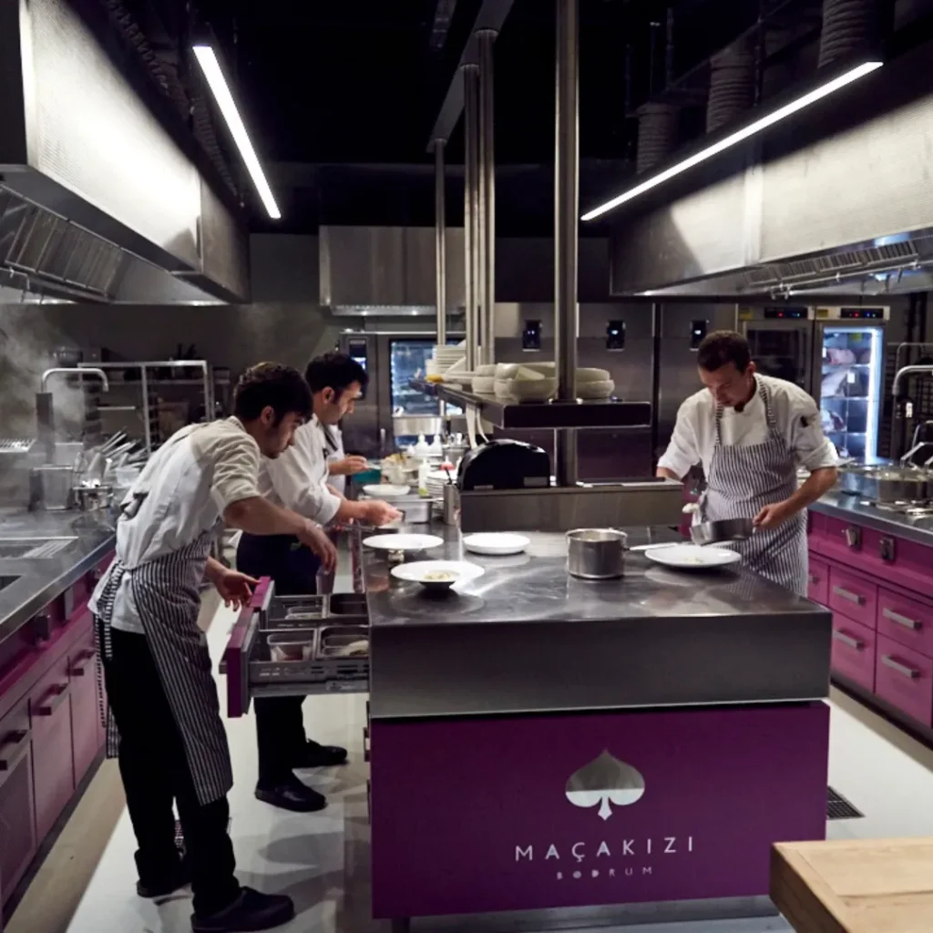 Equipo de chefs trabajando en una isla de cocina profesional con gabinetes morados y encimera de acero inoxidable, mientras preparan platos.