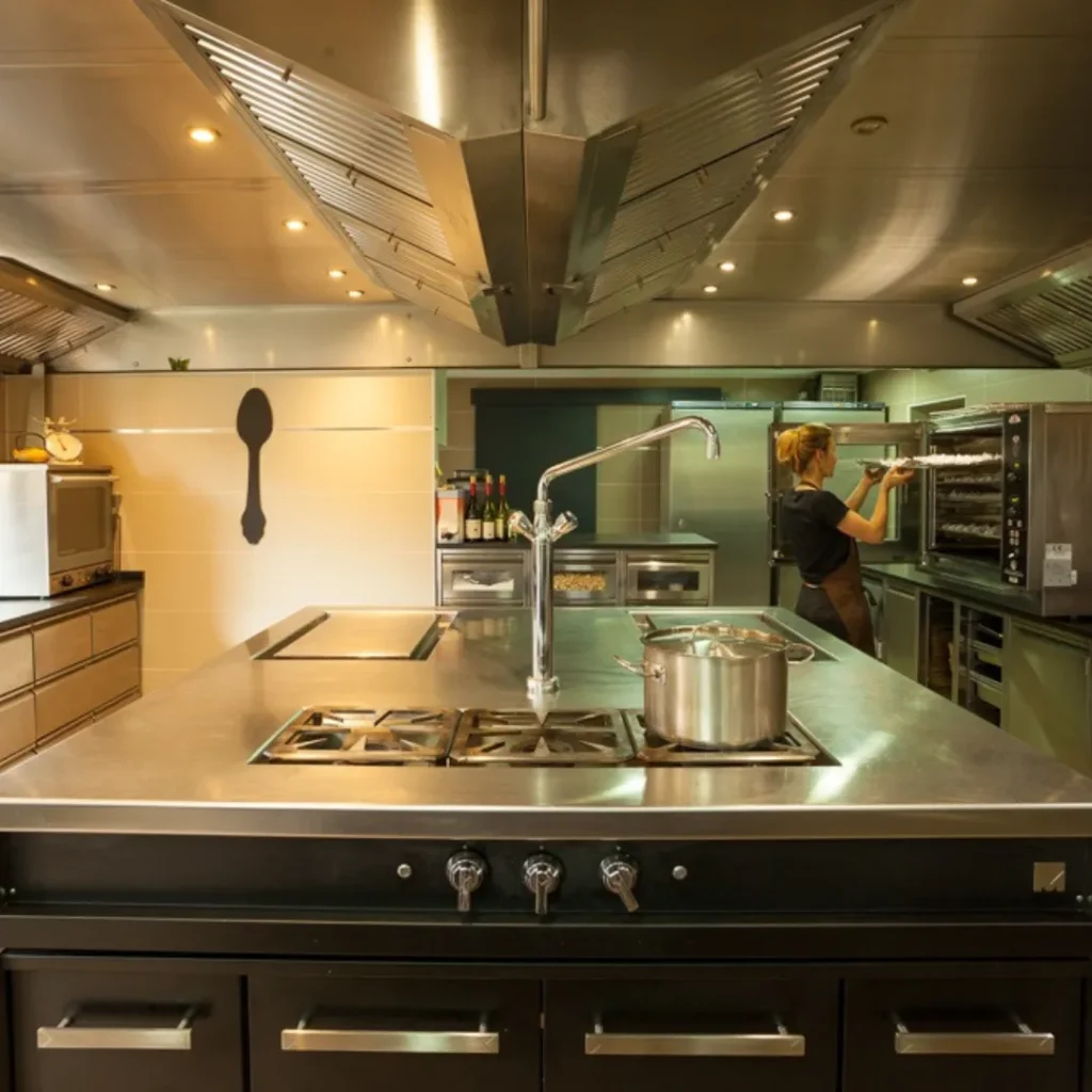 Isla central de acero inoxidable con zona de cocción y fregadero, en cocina industrial con horno y campana extractora.