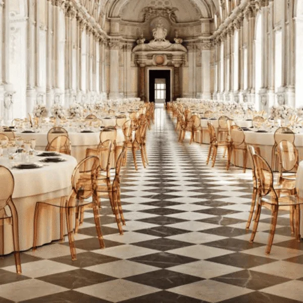 Salón de palacio decorado con mesas redondas y sillas translúcidas, ambiente de lujo para eventos exclusivos.